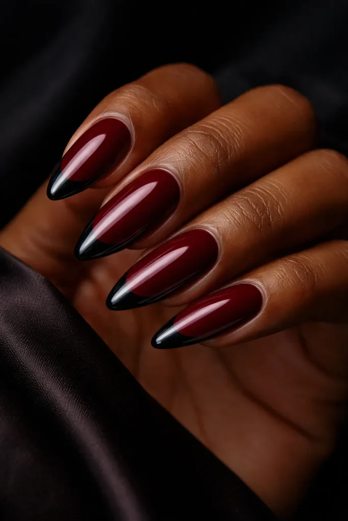Almond shaped burgundy nails with thin black French tips on deep melanin-rich skin.