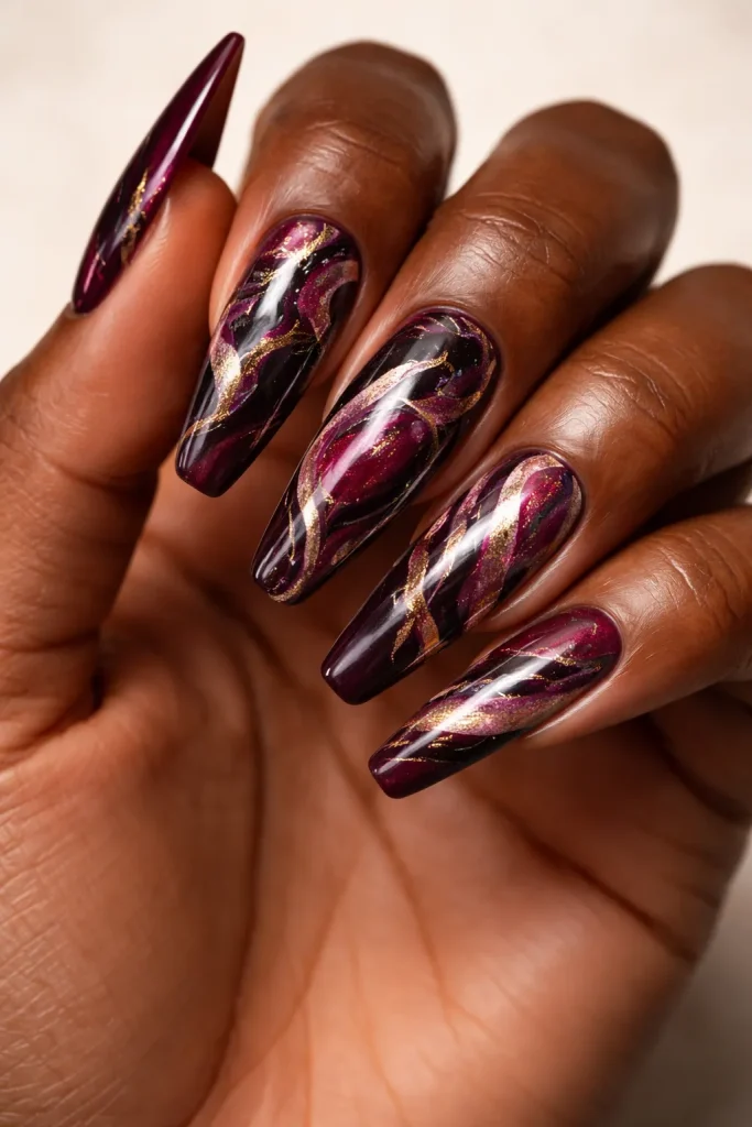 Intricate marble nail art using burgundy and gold swirls on a hand with mahogany skin.