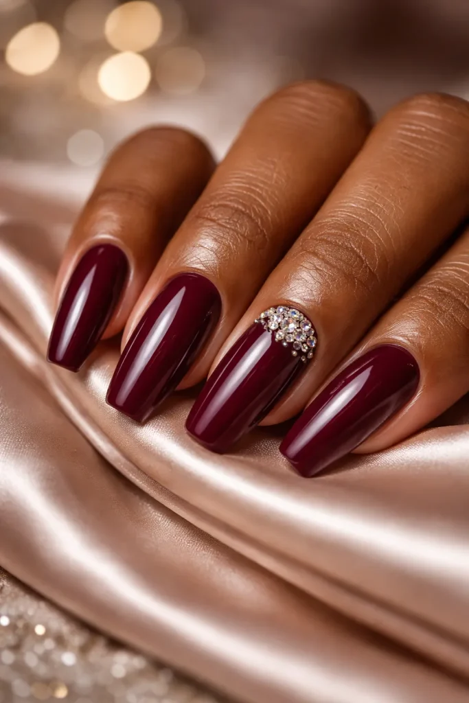 Deep burgundy nails with a cluster of small sparkling rhinestones on the ring finger.