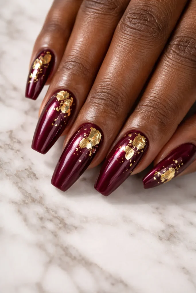 Burgundy nails decorated with gold foil accents on a hand with glowing chocolate skin.