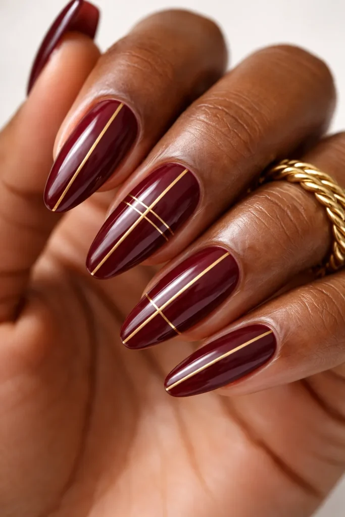 Thin geometric gold stripes on a deep burgundy nail base against cocoa-colored skin.