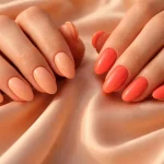 Hands featuring warm peach and vibrant coral nail polish against soft silk fabric.