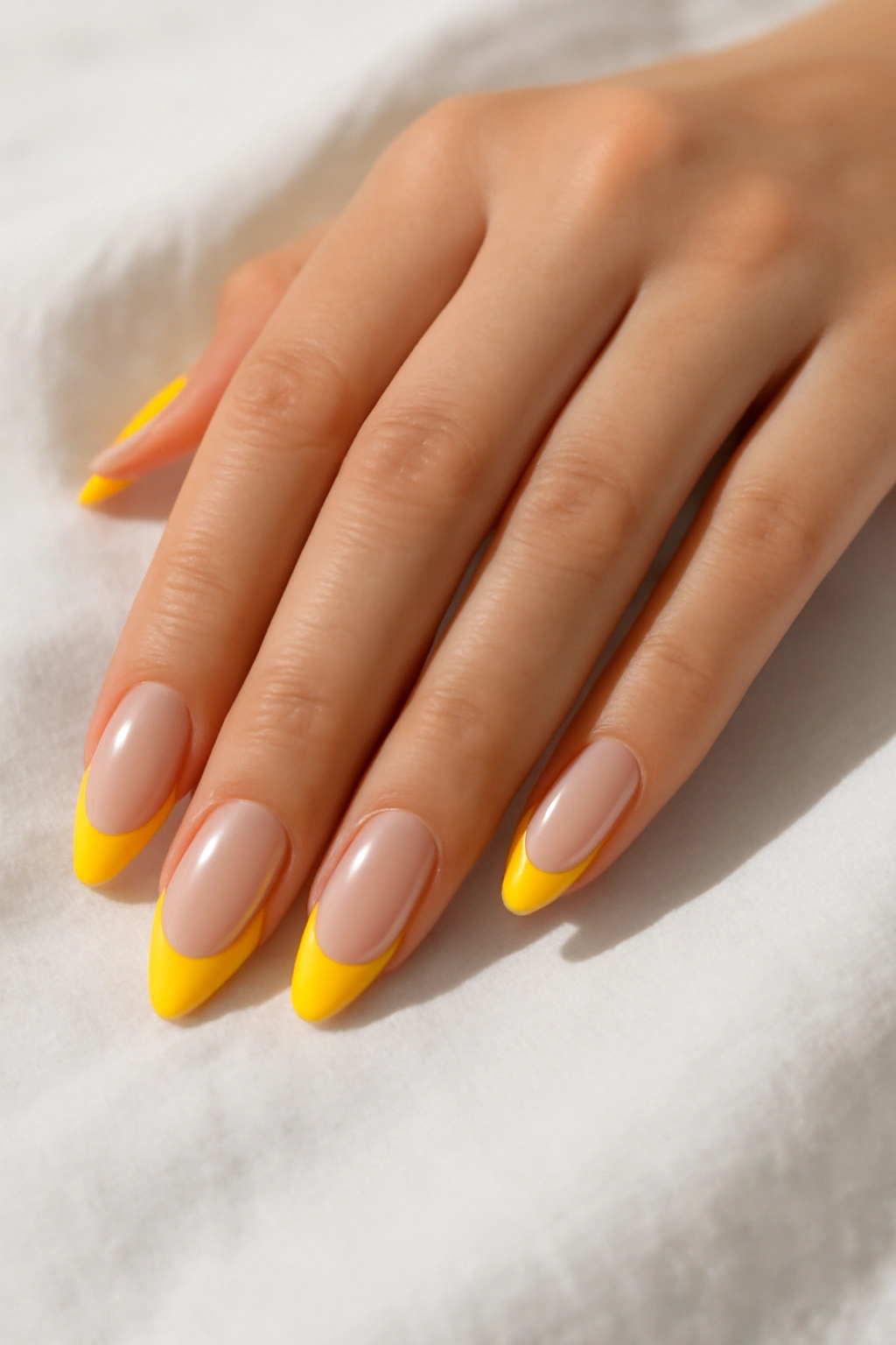 Close-up of almond nails with a clear base and bright yellow French tips on white linen.